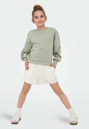 Ung pige står med benene spredte iført en lysegrøn sweatshirt med blomsterærmer og hvide shorts mod en ensfarvet baggrund.
