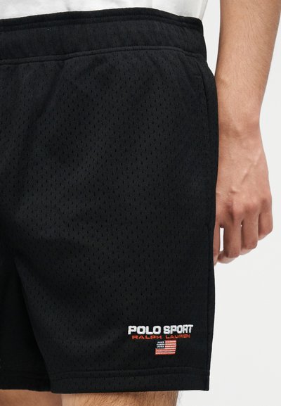 Μαύρα σορτς από διχτυωτό ύφασμα με λογότυπο "Polo Sport Ralph Lauren" και πάτς της αμερικανικής σημαίας στο κάτω μέρος, που φοριούνται από ένα άτομο που στέκεται.
