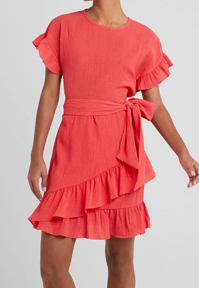 Krátké korálové šaty s volánkovými rukávy, vázáním v pase a vrstveným volánkovým lemem. Texturovaná tkanina dodává celkovému designu hloubku.