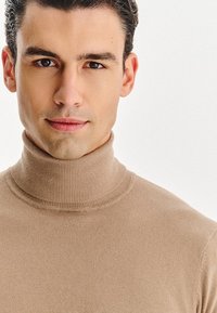 Beżowy sweter półgolf wykonany z dzianiny, z dopasowanym kołnierzem i gładką fakturą, noszony przez mężczyznę z krótkimi włosami i neutralnym wyrazem twarzy.