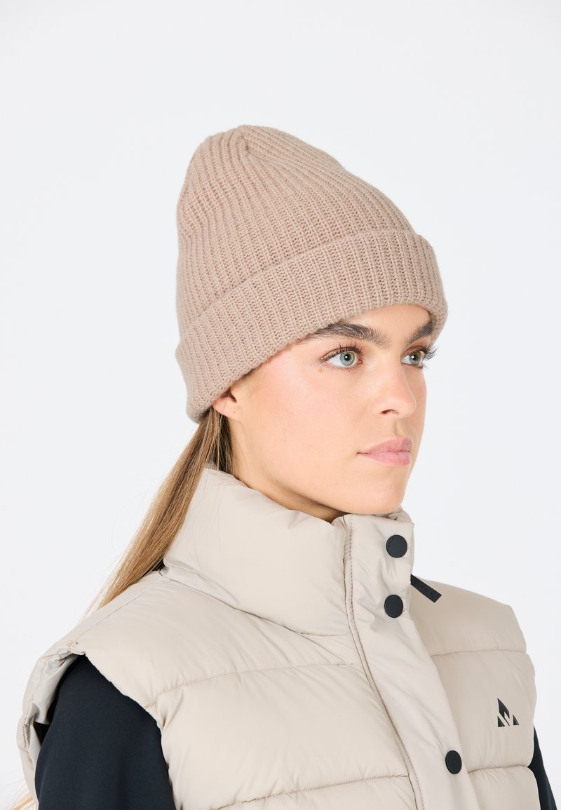 Beige stickad mössa med ribbad textur, med en rullad kant. Bärs tillsammans med en ljusfärgad puffer-väst och en svart jacka.