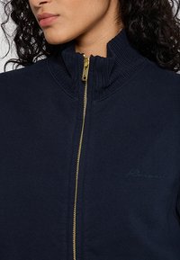 Veste zippée marine avec col montant, dotée d'une fermeture éclair dorée et d'un col côtelé. Logo brodé subtil sur la poitrine. Texture de tissu lisse.