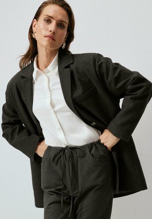 Blazer grigio scuro con rever a punta, indossato sopra una camicia bianca con bottoni. Abbinato a pantaloni con cordoncino in una tonalità grigia coordinata.