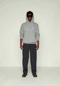 Uomo che indossa un hoodie grigio con logo circolare, pantaloni cargo neri, scarpe nere e grandi occhiali da sole neri, in piedi su uno sfondo semplice.