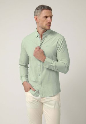 Hombre con camisa verde claro abotonada y pantalones blanco hueso, con una mano ajustando el cuello y la otra en el bolsillo, mirando hacia un lado.