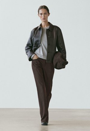 Femme marchant à l'intérieur, portant une veste en cuir marron foncé, une chemise grise, un pantalon marron, des chaussures noires, et tenant un sac en cuir marron assorti.