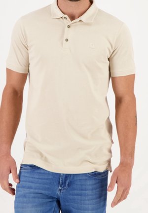 Homme portant un polo beige à manches courtes et un jean bleu, debout avec les bras détendus sur les côtés devant un fond blanc.
