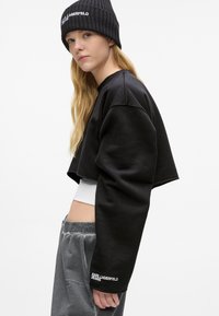 Črna skrajšana jakna iz mehkega blaga, z širokimi rokavi in vezenim logotipom. V kombinaciji s sivimi žametnimi trenirko in pleteno kapo.