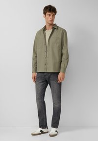 Man in een olijfgroene button-up shirt over een beige T-shirt, donkere grijze jeans en witte sneakers, staande tegen een effen lichtgrijze achtergrond.