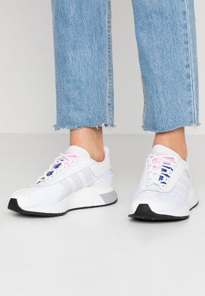 Scarpe da ginnastica bianche con sovrapposizioni testurizzate, tre strisce grigie, lacci rosa e blu, e suola in gomma nera. Pantaloni in denim con orlo sfilacciato.