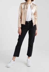 Metallisch rosegoldene Zip-Jacke mit hohem Kragen, getragen über einem weißen T-Shirt, kombiniert mit schwarzen knöchellangen Jeans und weißen Sneakern.
