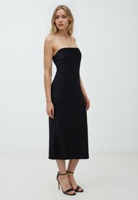 Schwarzes, trägerloses Midi-Kleid mit einem taillierten Oberteil und glatter Textur. Kombiniert mit schwarzen Riemchenabsätzen mit Knöchelriemen.