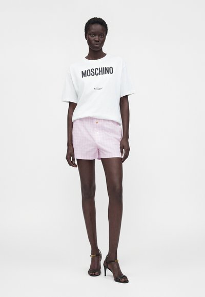 Femme debout portant un t-shirt blanc Moschino, un short rose clair et des sandales noires à talons hauts sur fond blanc uni.