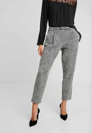 Femme portant un pantalon gris texturé taille haute avec une ceinture et des talons hauts pointus noirs, associé à un haut noir à manches longues avec un détail en dentelle.