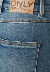 Jeans de mezclilla azul claro con una etiqueta de cuero marrón que dice "ONLY" y "Blue Denim". Costuras notables y un diseño en el bolsillo trasero.