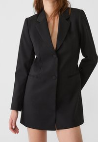 Abito blazer nero realizzato in tessuto liscio, caratterizzato da un colletto a punta, due bottoni, tasche laterali e una vestibilità sartoriale con un orlo più corto.