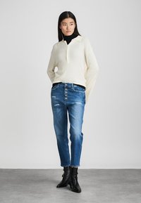 Cremefarbener, gerippter Strickpullover mit Kragen, kombiniert mit blauen, verwaschenen Jeans mit Knopfleiste und knöchelhohen schwarzen Lederstiefeln.