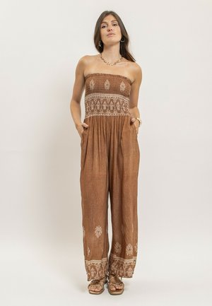 Femme debout les mains dans les poches, portant une combinaison marron sans bretelles avec des motifs blancs et des sandales beiges, sur un fond uni.