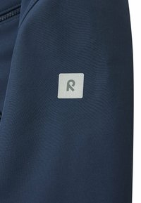 Nahaufnahme eines dunkelblauen Stoffes mit einem kleinen, quadratischen Logo, das ein stilisiertes "R" in Grau zeigt, um das Entwurfsdetail hervorzuheben.