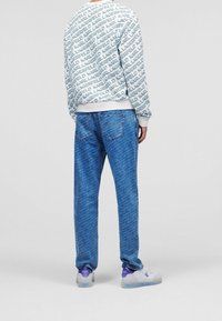 Felpa bianca con un motivo di testo blu ripetuto, abbinata a pantaloni di jeans blu con una leggera trama. Le calzature sono bianche con accenti viola.