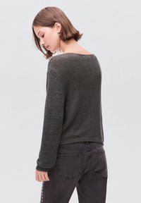 Junge Frau mit kurzen braunen Haaren, die einen dunkelgrauen Strickpullover und schwarze Jeans trägt, sich abwendet und nach unten nach links schaut.