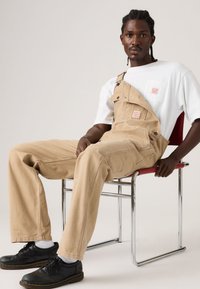 Άνδρας με πλεξούδες που φοράει μπεζ ολόσωμη φόρμα και λευκό μπλουζάκι, καθισμένος πλευρικά σε μια κόκκινη και χρωμιωμένη καρέκλα, κοιτάζοντας την κάμερα.