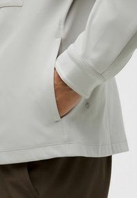 Helehall, tekstureeritud pika varrukaga pluus, millel on tasku ja peen logo detail, omades kerget laia lõiget ja mansetiga varrukaid.