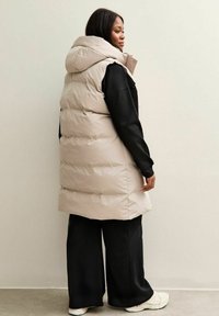 Chaleco acolchado beige claro con capucha, presenta un diseño acolchado y una forma ligeramente alargada, llevado sobre un suéter negro y pantalones de pierna ancha.