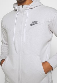 Světle šedá zipová mikina s vysokým límcem, bílými stahovacími šňůrkami a texturovaným logem Nike na hrudi. Obsahuje přední kapsy a žebrované manžety.