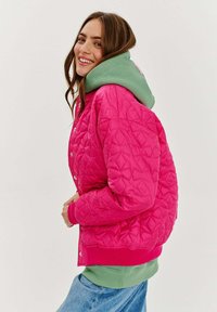 Ružová prešívaná bomber bunda s zelenou mikinou s kapucňou pod ňou. Obsahuje cvoky, rebrované manžety a geometrickú textúru.