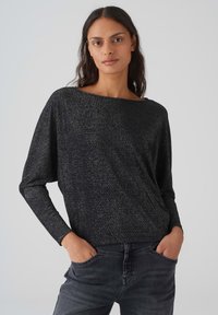 Lässiges langärmliges Oberteil in Schwarz mit metallischen Sprenkeln, Bootsausschnitt, strukturierter Stoff, kombiniert mit dunkelgrauen Jeans.