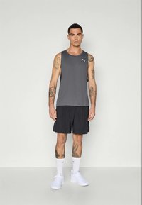 Mand med tatoveringer iført grå ærmeløs Puma-trøje, sorte shorts, hvide Puma-sokker og hvide sneakers, stående op ad en ensfarvet væg.