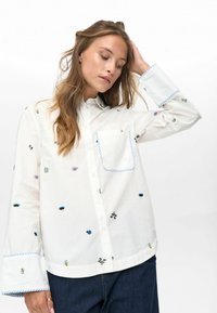Jonge vrouw met lang haar die een witte blouse met knopen en een blauw gestreepte rand draagt, voorzien van kleine kleurrijke geborduurde motieven, gecombineerd met donkere jeans.