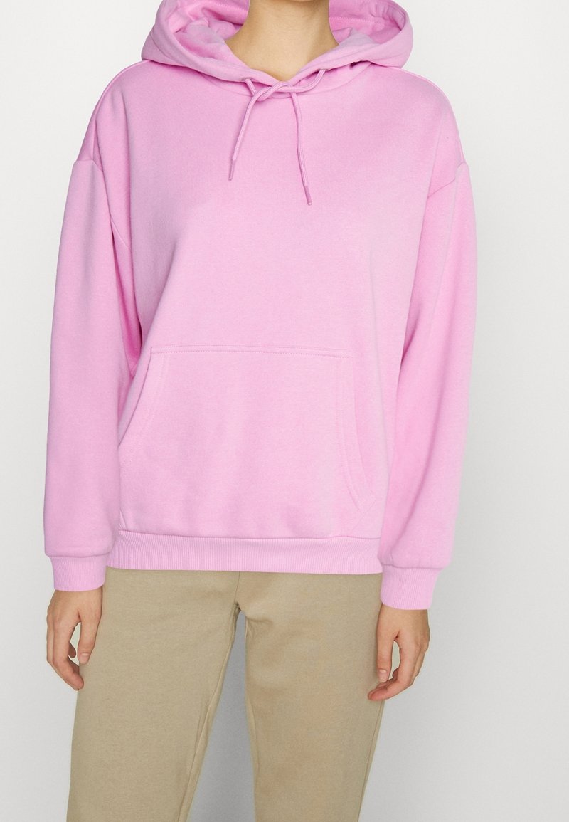 Personne portant un sweat à capuche rose clair avec poche avant et pantalon beige, debout devant un fond uni.