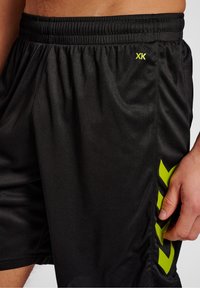 Svarta idrottsshorts med resår i midjan, med en texturerad tygkvalitet, samt en ljusgrön logotyp och designaccenter på sidan.