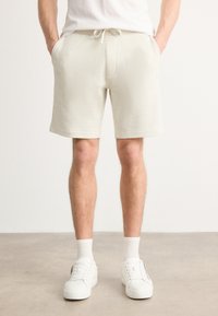 Shorts beige con texture, dotati di una fascia elastica in vita e coulisse. Il modello indossa sneakers bianche e calzini, in piedi su un pavimento neutro.