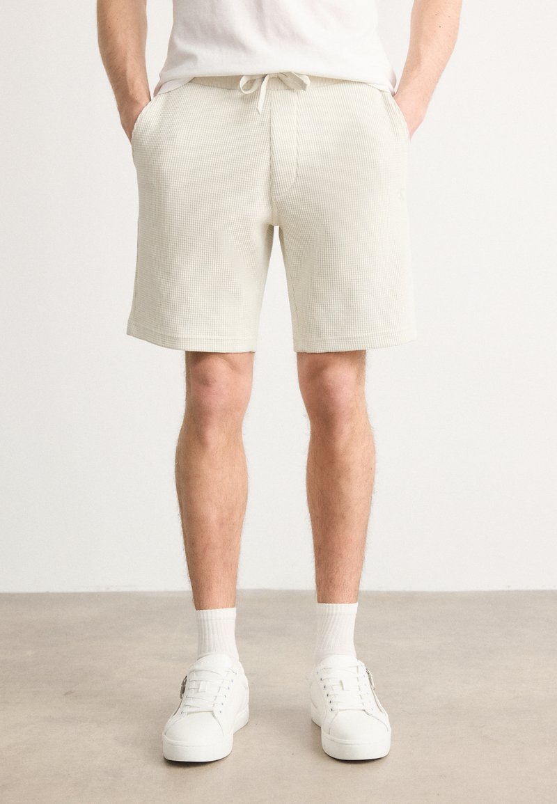 Shorts beige con texture, dotati di una fascia elastica in vita e coulisse. Il modello indossa sneakers bianche e calzini, in piedi su un pavimento neutro.