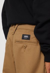 Khaki broek met een gladde textuur, voorzien van een knoopsluiting, achterzakken en een zwart Vans-label op de tailleband.