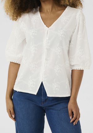 Woman wearing white floral embroidered buttoned blouse with puffed sleeves and blue jeans, standing against plain background.