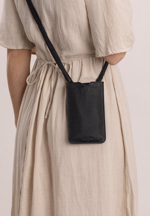 Svart läder crossbody mobilväska med pebblad textur, med en rundad övre kant och en tunn rem, bärs mot en ljusbeige klänning.