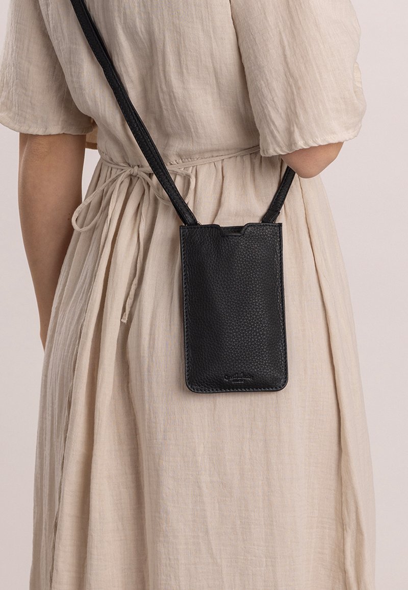 Černá kožená crossbody taška na telefon s jemnou texturou, se zaobleným horním okrajem a tenkým popruhem, nošená přes světle béžové šaty.