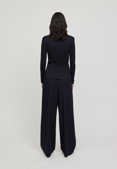 Crna majica dugih rukava od glatkog materijala, u kombinaciji s crnim hlačama širokih nogavica. Outfit je jednostavan, bez vidljivih uzoraka ili naglasaka.