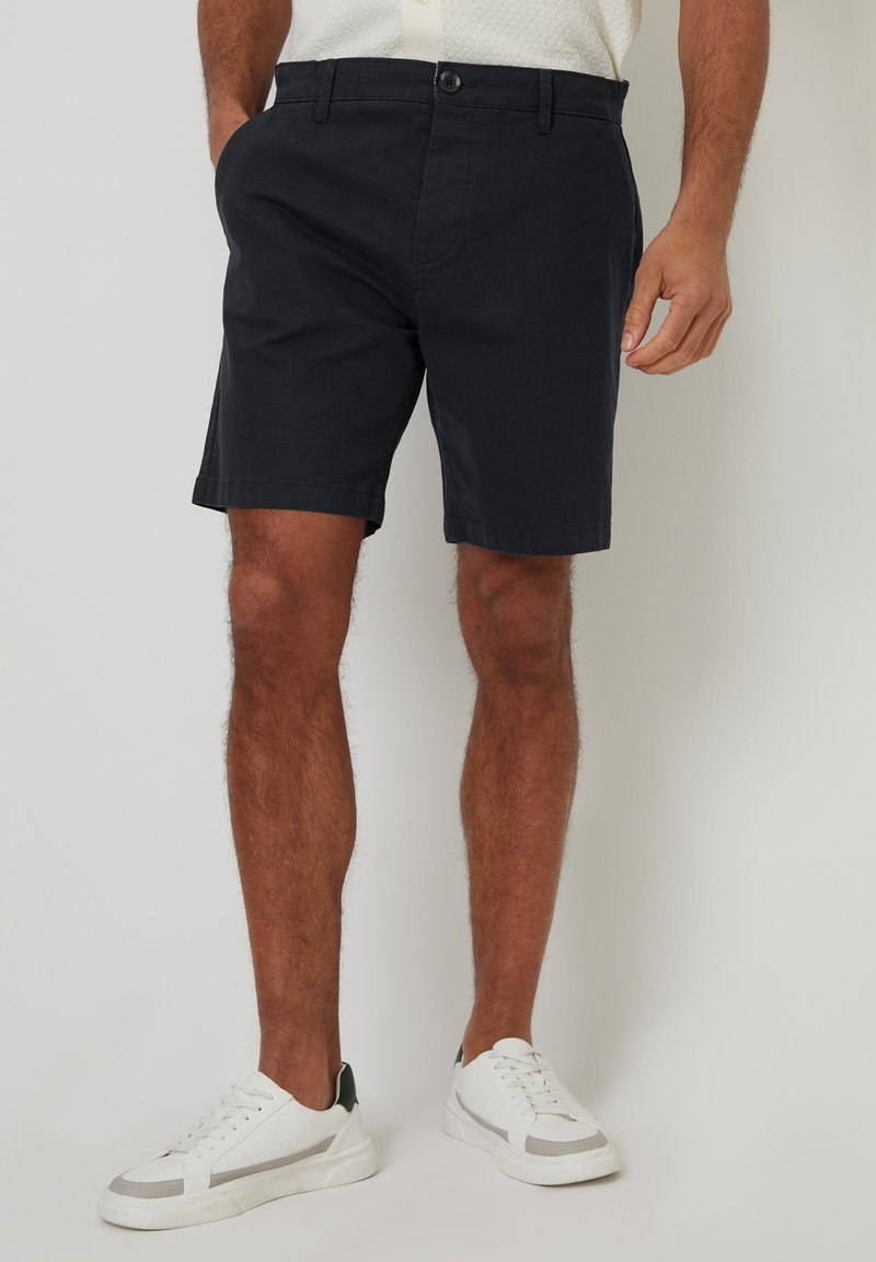 Marineblauwe shorts van een katoenen mixstof met een platte voorkant, zijzakken en een enkele knoopsluiting, gecombineerd met witte sneakers.