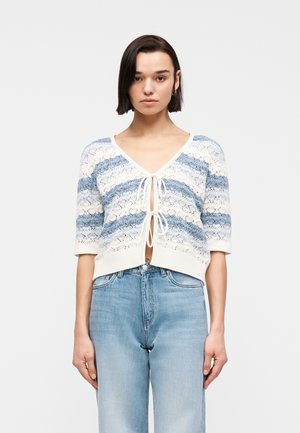 Γυναίκα που φοράει πλεκτή ζακέτα με μπλε και άσπρες ρίγες, δεμένη με δύο φιόγκους μπροστά, και ανοιχτόχρωμο τζιν ψηλόμεσο παντελόνι, σε λευκό φόντο.