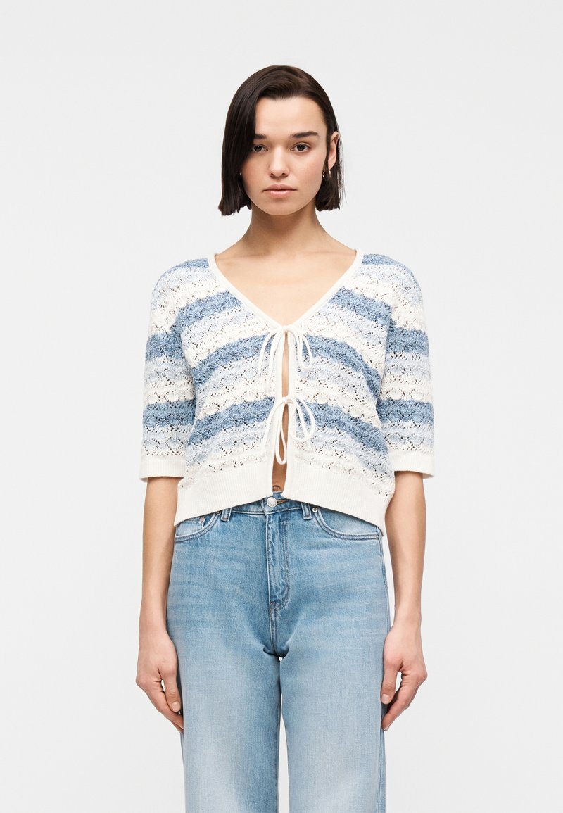 Γυναίκα που φοράει πλεκτή ζακέτα με μπλε και άσπρες ρίγες, δεμένη με δύο φιόγκους μπροστά, και ανοιχτόχρωμο τζιν ψηλόμεσο παντελόνι, σε λευκό φόντο.