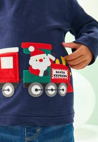 Dijete nosi tamnoplavu majicu s izvezbenim Djedom Božićnjakom koji maše iz crveno-zelene lokomotive označene s "Santa Express".