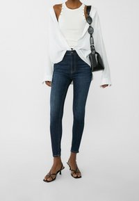 Camisa blanca de botones sobre un top de tirantes blanco ajustado, combinada con jeans skinny oscuros y tacones negros con tiras. Bolso negro con una correa texturizada.