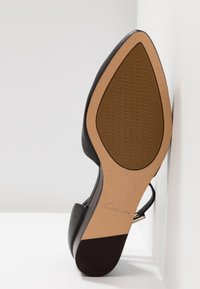Chaussure à talon noir avec une tige en cuir lisse, un bout pointu et une semelle texturée beige. Comprend une fine bride de cheville et des accents polis.