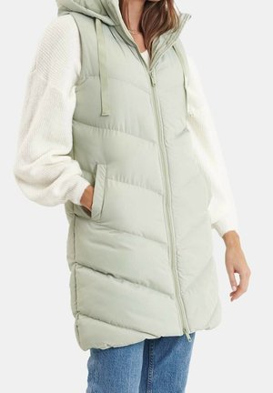 Femme portant un gilet matelassé sans manches à capuche vert clair sur un pull blanc et un jean bleu, avec une main dans la poche, devant un fond uni.