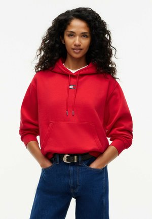 Mujer con cabello rizado lleva sudadera roja con bolsillo delantero y vaqueros azules, de pie con las manos en los bolsillos frente a fondo blanco.